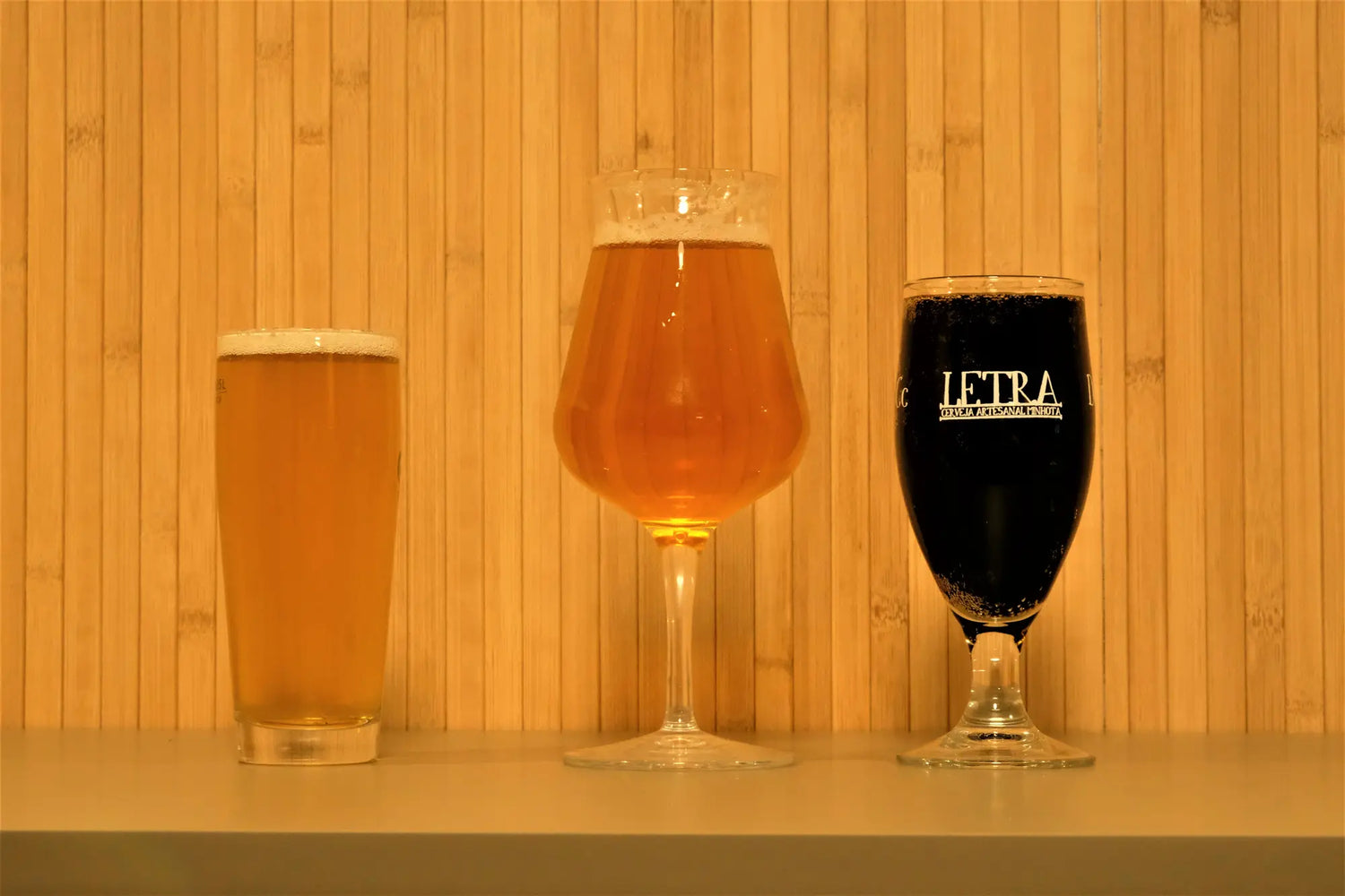 Três copos de cheios com cerveja artesanal, um copo de cerveja pilsner à esquerda e mais pequeno que os outros, o do meio em copo tulipa com cerveja dourada nele e terceiro copo à direita com uma stout e com inscrição da marca letra no vidro. Fundo com ripado de madeira