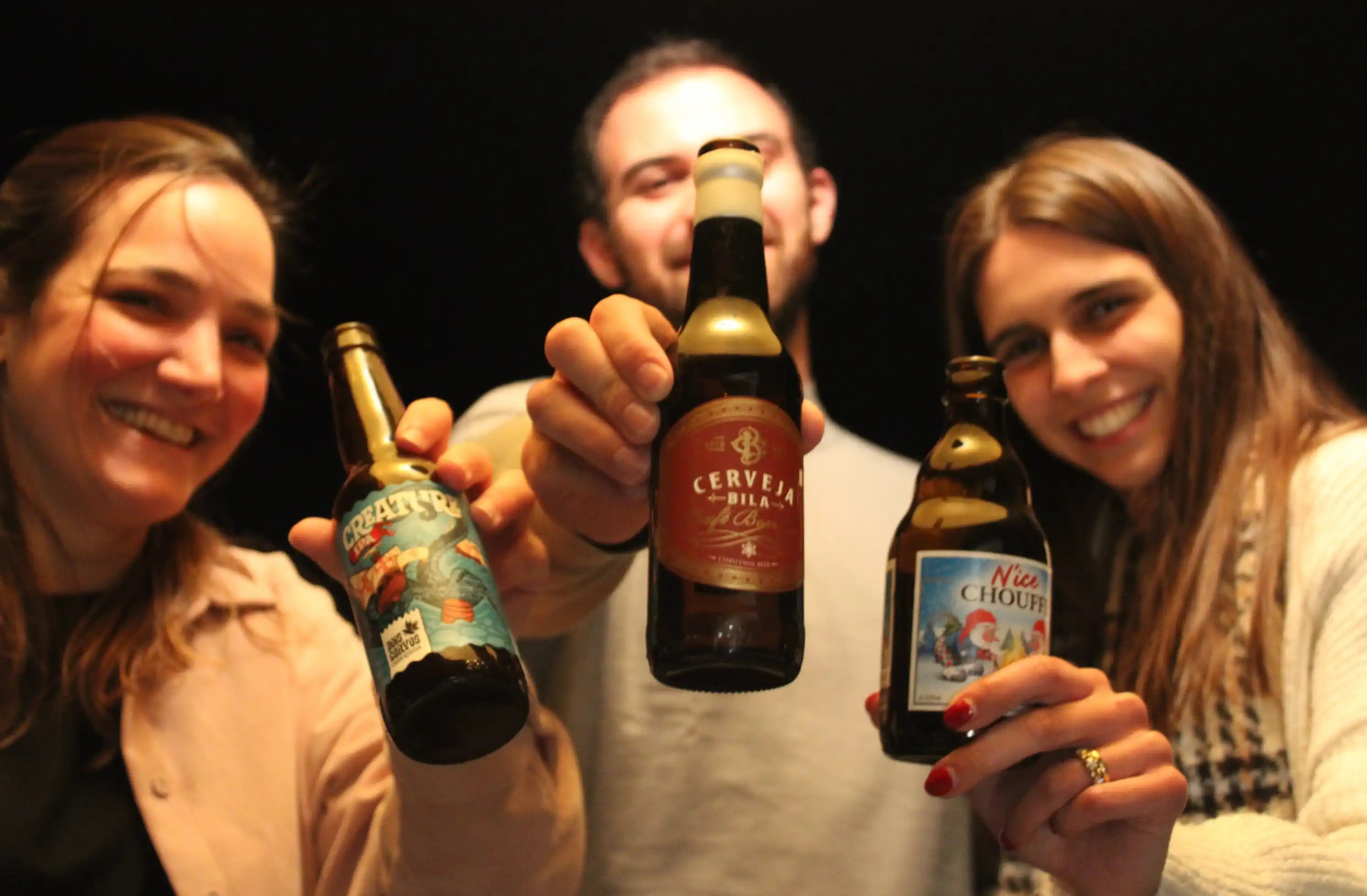 Duas raparigas e um rapaz a brindarem com três cervejas artesanais num ambiente divertido e fundo escuro
